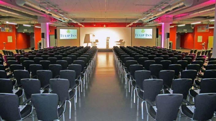Konferenzraum mit Reihenbestuhlung und Bühne im Tulip Inn Hotel Düsseldorf Arena. Auf der Bühne stehen ein Rednerpult, ein Tisch und zwei Stühle. Links und rechts zeigen Bildschirme das Tulip Inn-Logo. Der Raum ist in Rot- und Grautönen gehalten und mit Deckenbeleuchtung sowie Belüftungssystemen ausgestattet.