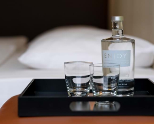 Tulip Inn Düsseldorf Arena Tablett mit einer Flasche Wasser mit dem Etikett „ENJOY“ und zwei Gläsern auf einer lederbezogenen Sitzfläche, im Hintergrund ein Hotelbett mit weißer Bettwäsche.