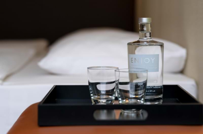 Tablett mit einer Flasche Wasser mit dem Etikett „ENJOY“ und zwei Gläsern auf einer lederbezogenen Sitzfläche, im Hintergrund ein Hotelbett mit weißer Bettwäsche.