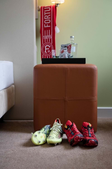 Hotelzimmerausschnitt mit einem braunen Hocker, auf dem ein Tablett mit Wasserflasche, Gläsern und Flyer steht. An der Wand hängt ein rot-weißer Fortuna-Düsseldorf-Schal unter einer Lampe. Auf dem Boden davor stehen ein Paar gelbe Fußballschuhe und ein Paar rote Sportschuhe.