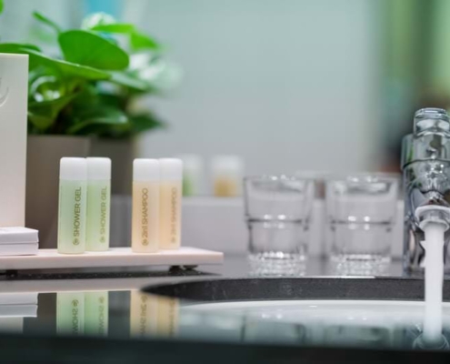 Tulip Inn Düsseldorf Arena Waschbecken im Hotelbad mit laufendem Wasserhahn, zwei Gläsern, einem Pflanzentopf und einem Set mit kleinen Fläschchen Duschgel und 2-in-1-Shampoo auf einem Tablett.