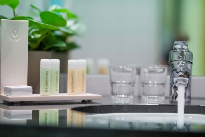 Waschbecken im Hotelbad mit laufendem Wasserhahn, zwei Gläsern, einem Pflanzentopf und einem Set mit kleinen Fläschchen Duschgel und 2-in-1-Shampoo auf einem Tablett.
