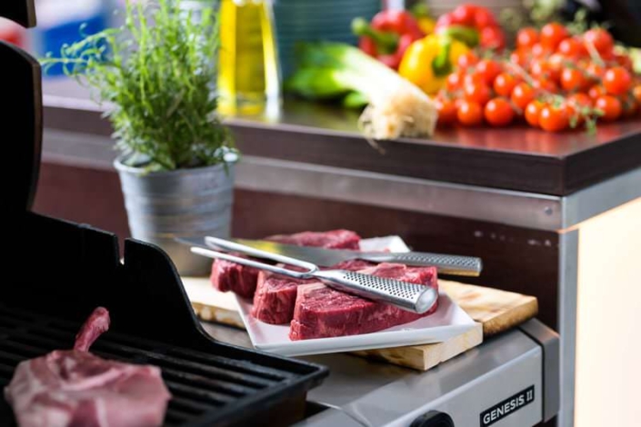 Nahaufnahme eines Grills mit rohem Fleischstück auf dem Rost. Daneben ein Teller mit mehreren Rinderfilets und Grillbesteck auf einem Schneidebrett. Im Hintergrund frisches Gemüse, Kräuter und eine Flasche Öl auf einer Küchenzeile.