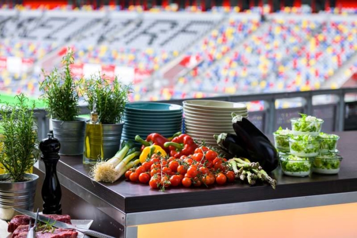 Zubereitungstisch mit frischem Gemüse, darunter Tomaten, Lauch, Paprika, Auberginen, Spargel sowie Kräutertöpfen und kleinen Salatschalen. Daneben Tellerstapel und Ölflaschen. Im Hintergrund unscharf die Ränge der MERKUR SPIEL-ARENA Düsseldorf.