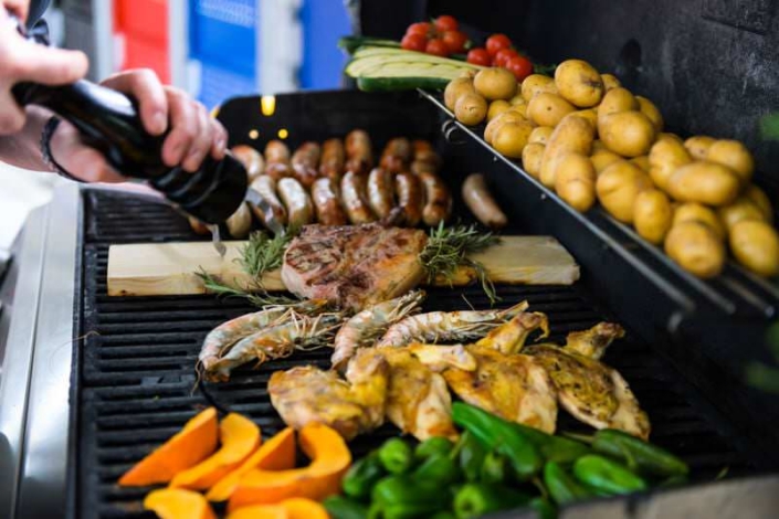 Grillbuffet mit frischen Zutaten wie Tomaten, Spargel, Kräutern, Brot, Nudeln, Salat und rohem Fleisch auf einer Terrasse mit Blick ins Stadion. Links ein geöffneter Grill mit Kartoffeln und Gemüse, im Hintergrund die leeren Ränge der MERKUR SPIEL-ARENA Düsseldorf.