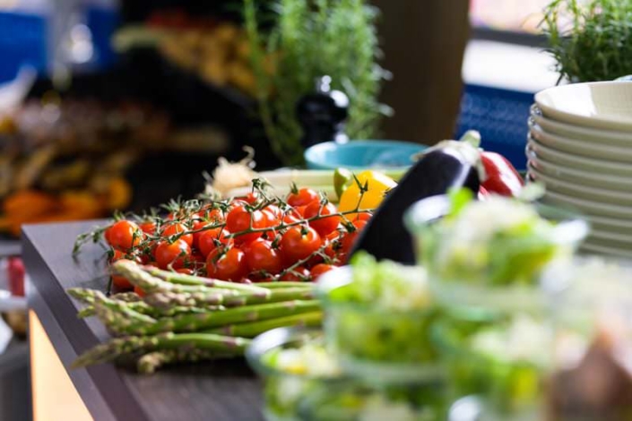 Frisches Gemüse auf einem Buffet-Tisch, darunter Kirschtomaten an der Rispe, grüner Spargel, eine Aubergine, Zitronen und kleine Schalen mit Salat. Im Hintergrund Tellerstapel, Kräuter und unscharf weitere Speisen.