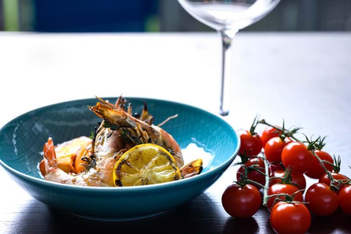 Tiefblaue Schale mit gegrillten Garnelen, Zitronenscheiben und Kräutern auf einem dunklen Holztisch. Daneben liegt eine Rispe mit frischen Cocktailtomaten, im Hintergrund ein leerer Weinkelch.