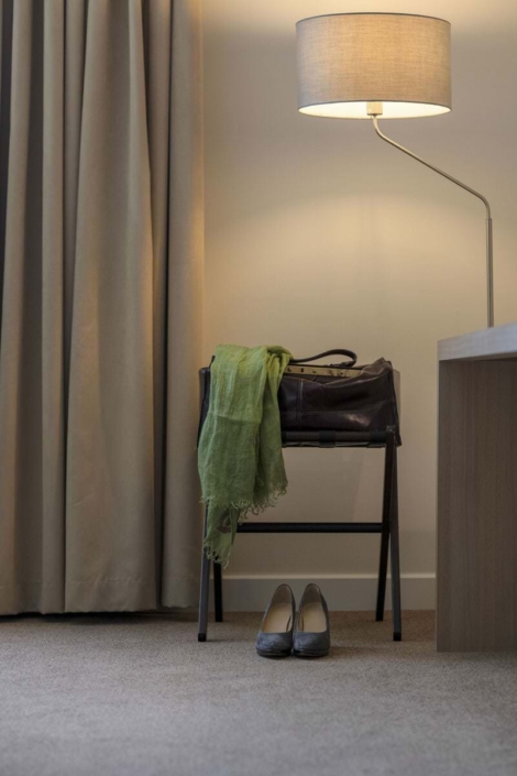 Hotelzimmerecke mit beigem Vorhang, dunkler Lederreisetasche auf einem Kofferbock, darüber ein grüner Schal. Auf dem Boden steht ein Paar graue Damenschuhe.