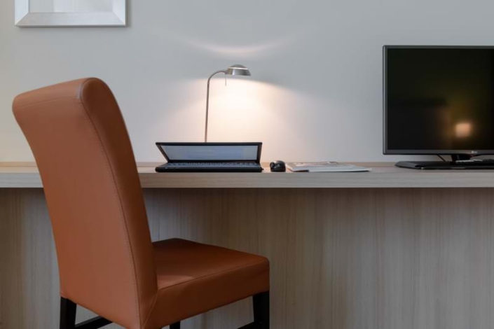 Schreibtisch mit einem Lederstuhl davor. Auf dem Schreibtisch ein Laptop und ein Fernseher.