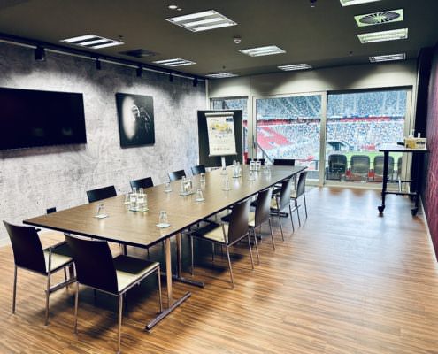 Event Loge 50 m² Block Ein moderner Konferenzraum mit langem Tisch, mehreren Stühlen, einer Wand mit großem Bildschirm und einem gerahmten Bild. Der Raum hat eine große Fensterfront mit Blick auf ein Stadion.