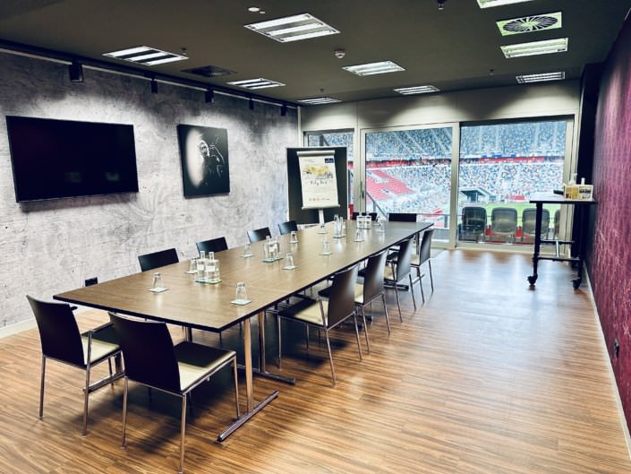 Ein moderner Konferenzraum mit langem Tisch, mehreren Stühlen, einer Wand mit großem Bildschirm und einem gerahmten Bild. Der Raum hat eine große Fensterfront mit Blick auf ein Stadion.