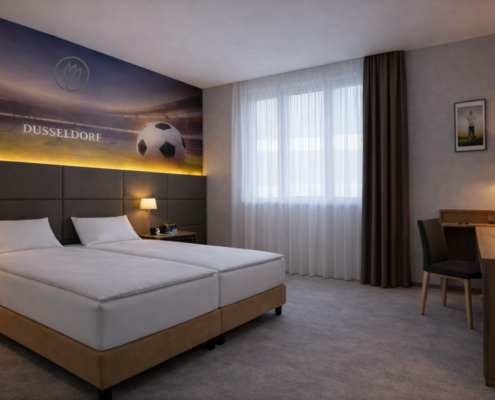 Zimmer 2026 Hotelzimmer mit zwei Einzelbetten, einem Schreibtisch mit Stuhl und Lampe, einem Flachbildfernseher auf einem Sideboard mit Minibar sowie einem Fenster mit Vorhang und einem kleinen Sitzbereich.