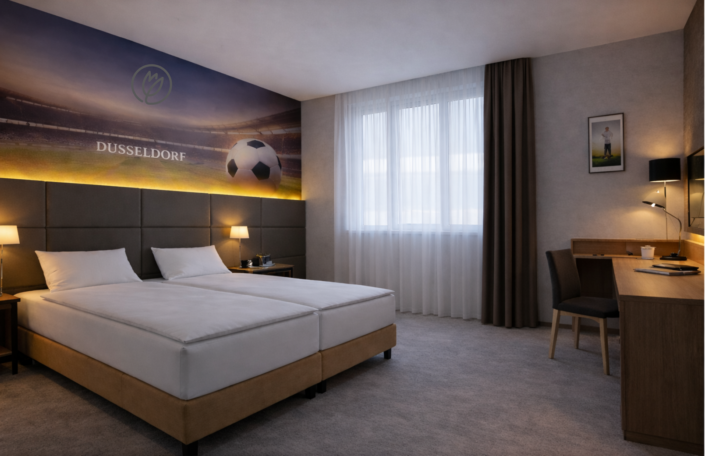 Hotelzimmer mit zwei Einzelbetten, einem Schreibtisch mit Stuhl und Lampe, einem Flachbildfernseher auf einem Sideboard mit Minibar sowie einem Fenster mit Vorhang und einem kleinen Sitzbereich.