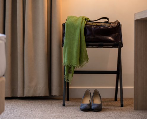 Zimmer Koffer Schal_2 Hotelzimmerecke mit beigem Vorhang, dunkler Lederreisetasche auf einem Kofferbock, darüber ein grüner Schal. Auf dem Boden steht ein Paar graue Damenschuhe.