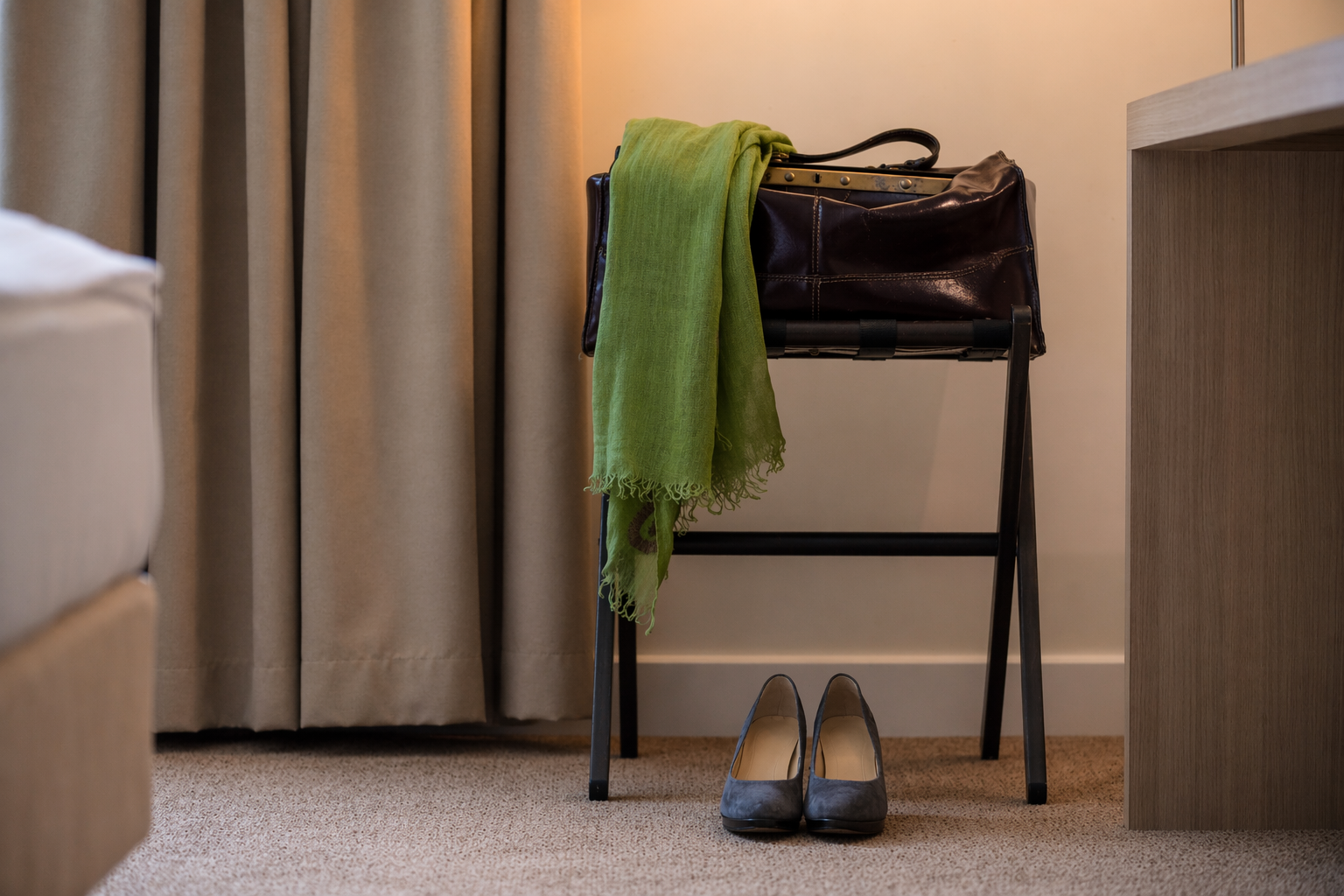 Zimmer Koffer Schal_2 Hotelzimmerecke mit beigem Vorhang, dunkler Lederreisetasche auf einem Kofferbock, darüber ein grüner Schal. Auf dem Boden steht ein Paar graue Damenschuhe.