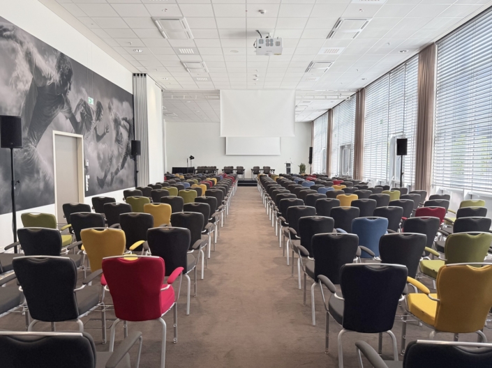 Tagungsraum in Reihenbestuhlung mit Mittelgang. Die meisten Stühle sind anthrazitfarben, einzelne haben rote, gelbe, grüne oder blaue Bezüge. Vorn stehen ein Podium mit einer Tischreihe, ein Rednerpult und eine große Projektionsleinwand. An der Decke hängt ein Beamer, seitlich stehen Lautsprecher auf Stativen. Links zeigt ein großes Schwarz-Weiß-Wandbild laufende Fußballspieler, rechts ist eine bodentiefe Fensterfront mit Jalousien und Vorhängen
