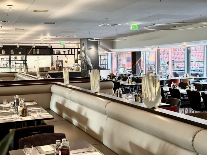Restaurant mit langer, beigefarbener Lederbank, die den Raum teilt; darauf stehen schmale Leuchtobjekte. Die Tische sind mit Platzsets, Gläsern, Besteck und Gewürzen eingedeckt, dazu dunkle Polsterstühle. Im Hintergrund eine Buffetzeile mit schwarzem Regalsystem und ein großes Schwarz-Weiß-Wandbild eines Sportlers. An der Decke hängen runde Lichtringe. Durch die Fensterfront rechts fällt der Blick auf die roten Sitzreihen des Stadions.