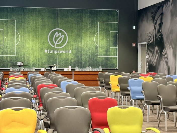 Der Blick geht über bunte Stuhlreihen in Grau, Rot, Gelb, Blau und Hellgrün auf die Stirnwand eines Tagungsraums. Dort zeigt ein Wandbild ein Fußballfeld mit Tulpen-Logo und dem Schriftzug „#tulipsworld“. Davor stehen ein Holzsideboard mit Wasserflaschen und Gläsern sowie zwei Stehtische. Rechts zeigt ein großes Schwarz-Weiß-Wandbild eine laufende Fußballspielerin.